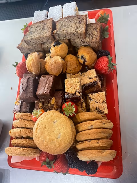 Red tray filled with an assortment of baked goods including cookies, brownies, bread slices, muffins, fruit bars, strawberries, and blackberries.