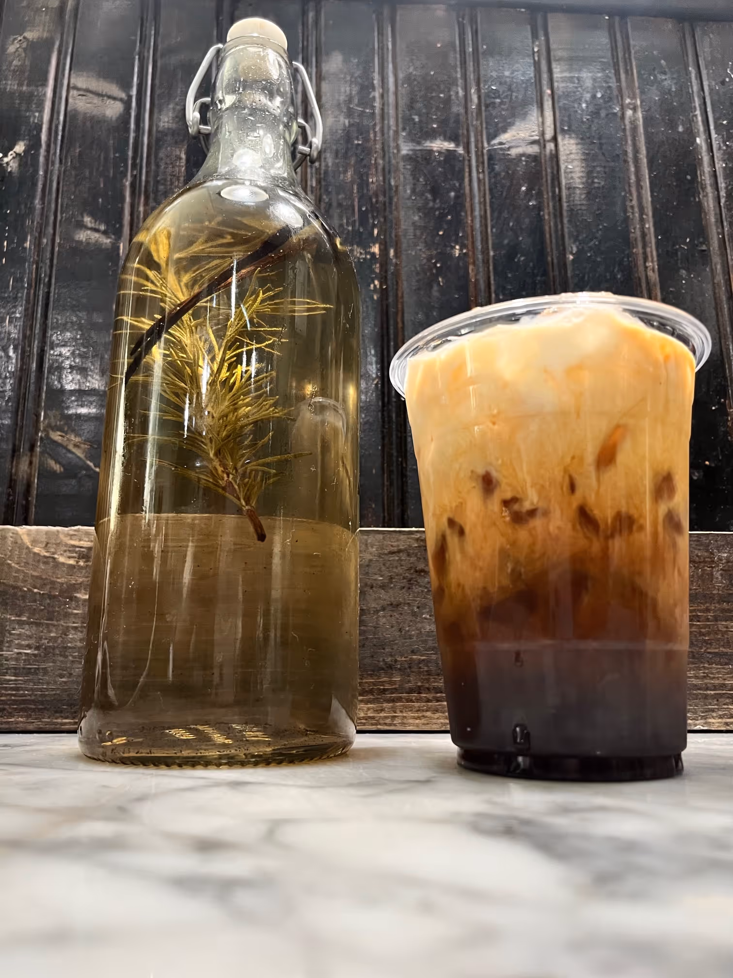 Glass bottle with a sprig of herb inside next to a plastic cup of iced coffee with cream on a marble surface.
