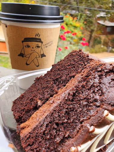 Close-up of two slices of moist chocolate cake in a clear plastic container with a paper coffee cup in the background featuring a cartoon face and the text 'Careful, I'm hot.'