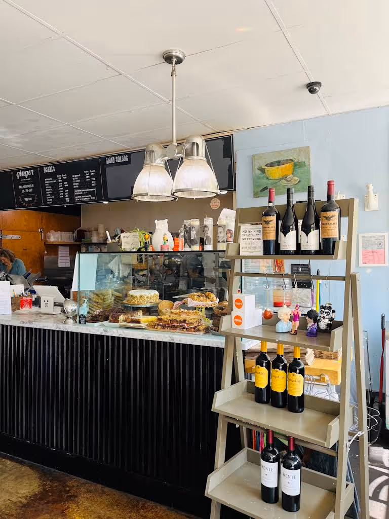 Interior of a cafe with a counter displaying pastries under a glass case and a shelf holding bottles of wine and small decorative items.