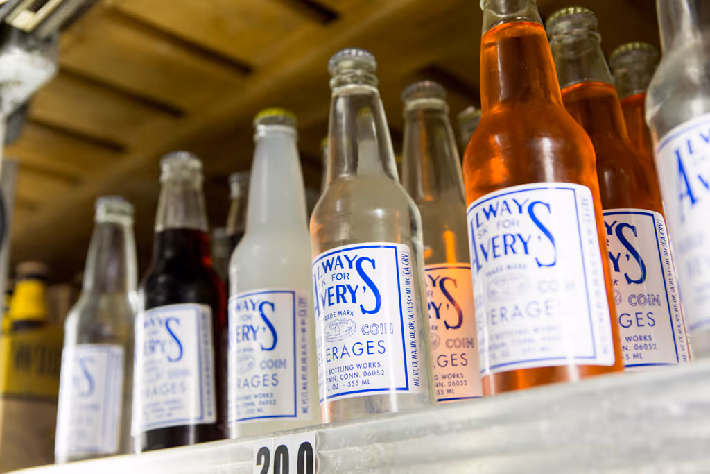 Row of vintage soda bottles with blue and white labels on a wooden shelf.