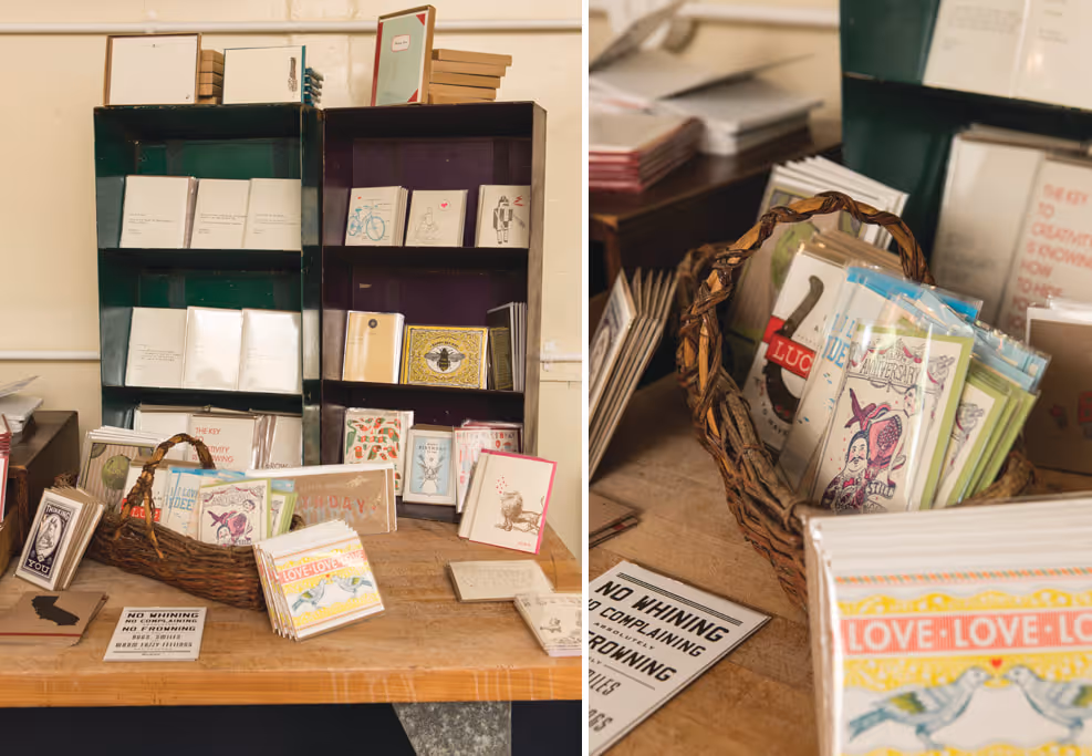 Wooden table and shelves displaying various small notebooks and cards, including a basket holding colorful illustrated cards and a sign reading 'No whining, no complaining, no frowning.'