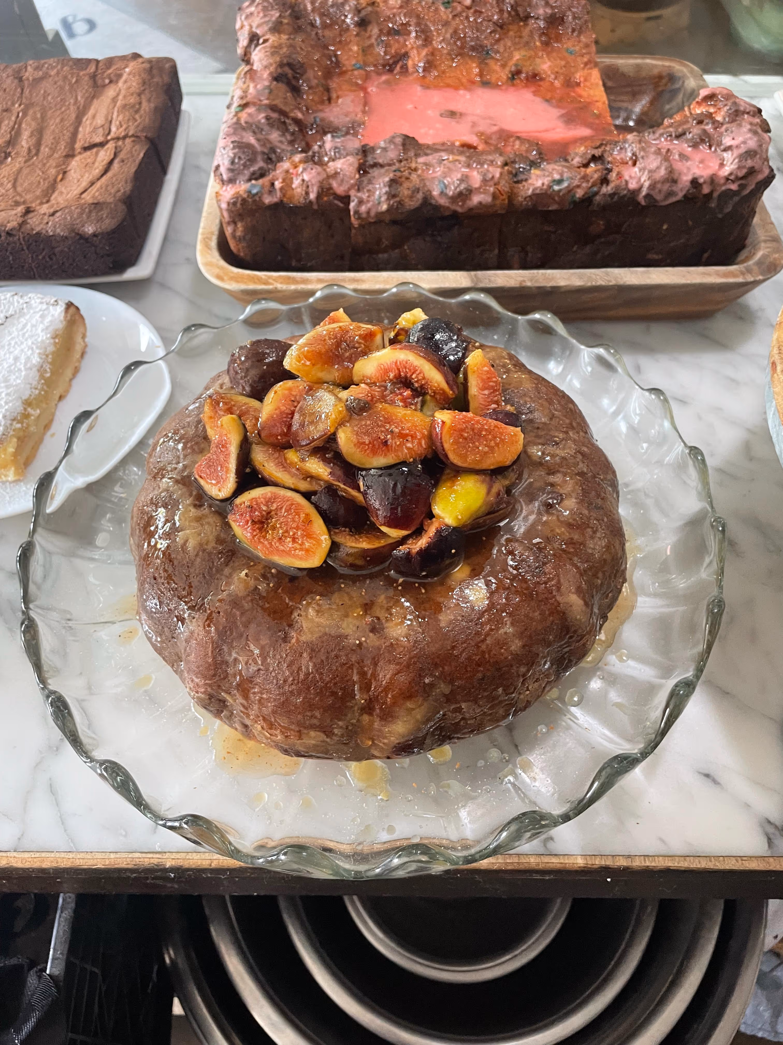 A glass plate with a glazed bundt cake topped with sliced figs, surrounded by other desserts on a marble countertop.