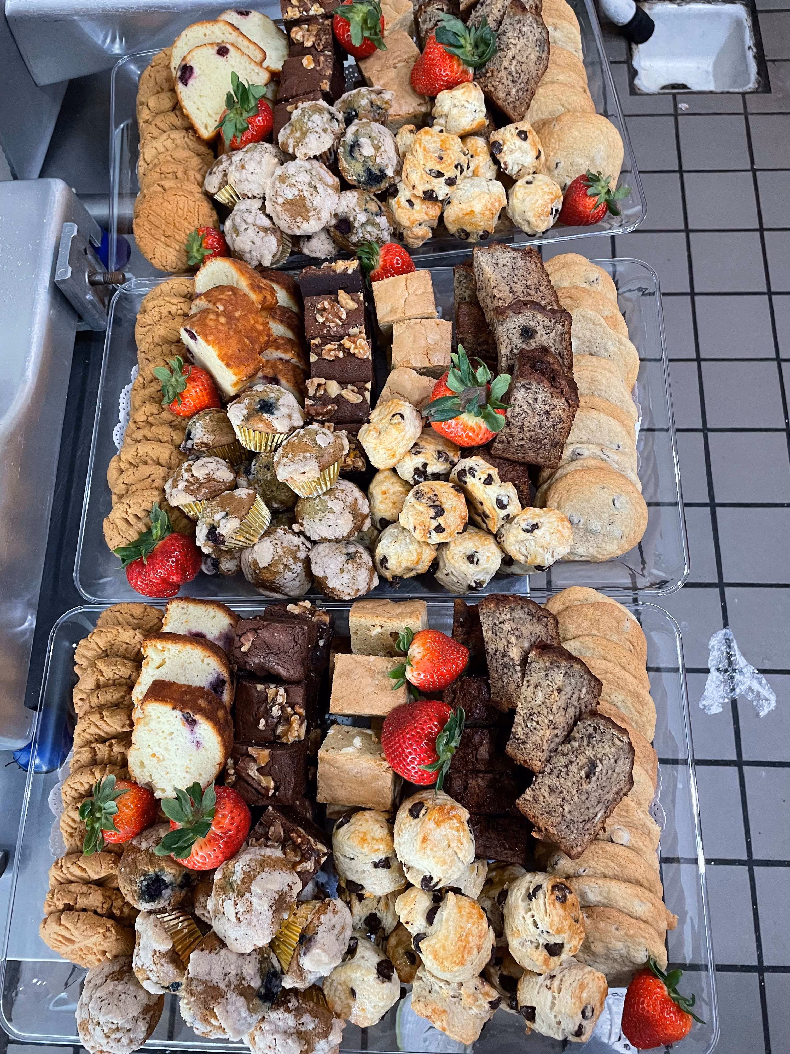 Three trays with an assortment of cookies, brownies, scones, muffins, banana bread slices, and strawberries arranged on a tiled kitchen counter.