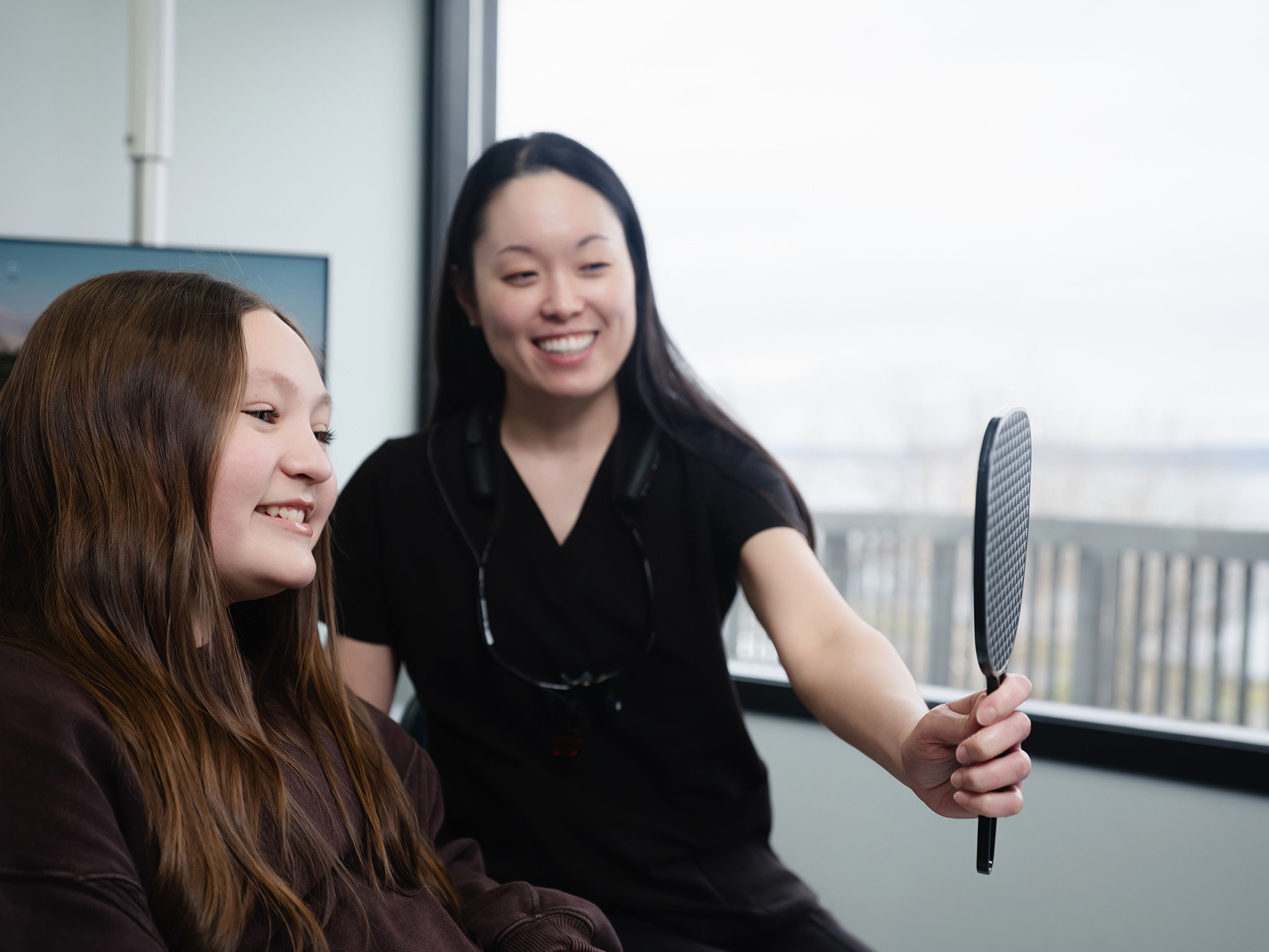 Smiling dental professional showing a young female patient her teeth using a handheld mirror.