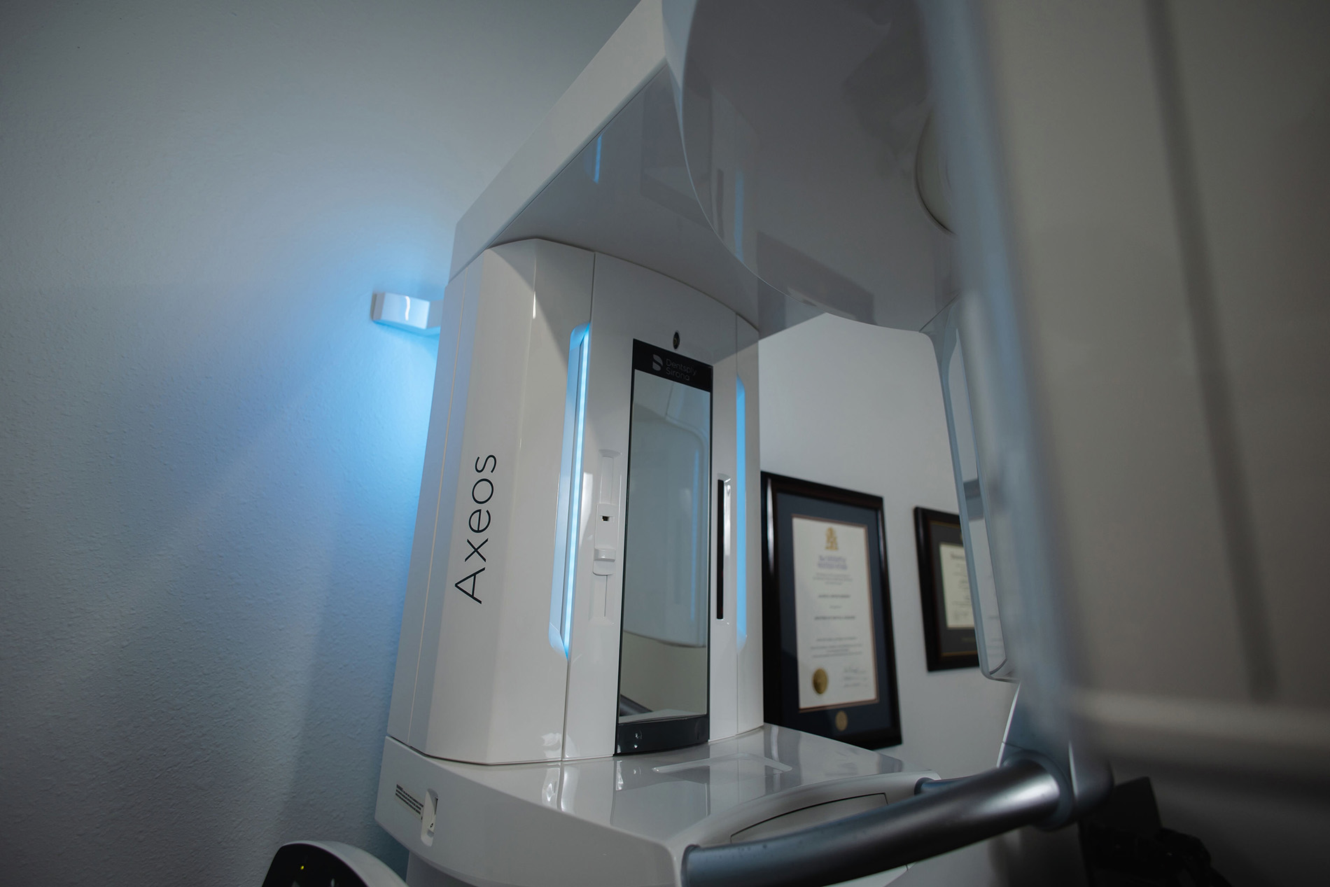Modern white Axceos dental imaging machine inside a clinic room with certificates on the wall.