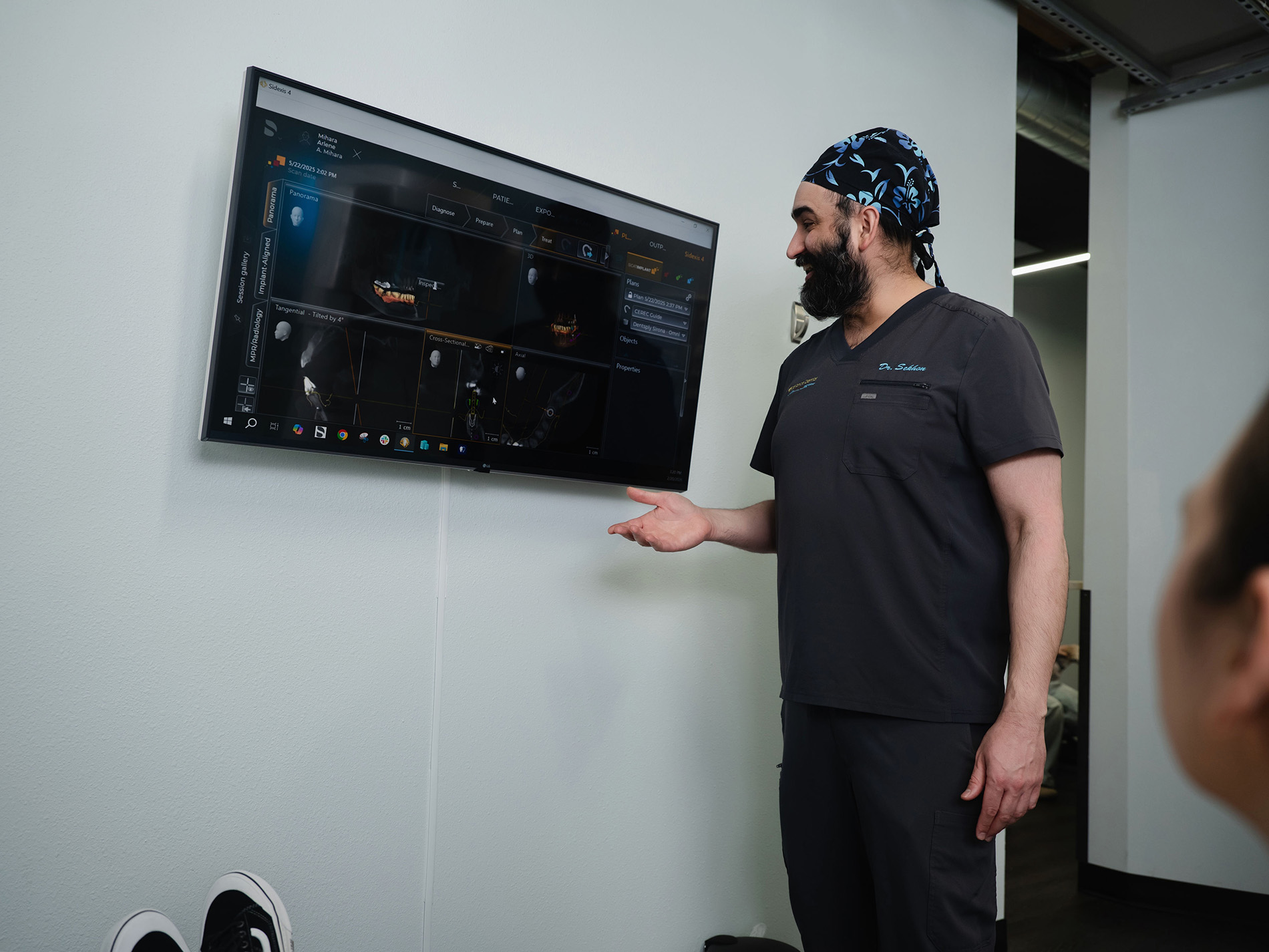 Dentist wearing scrub cap explaining dental x-ray images displayed on a wall-mounted monitor.
