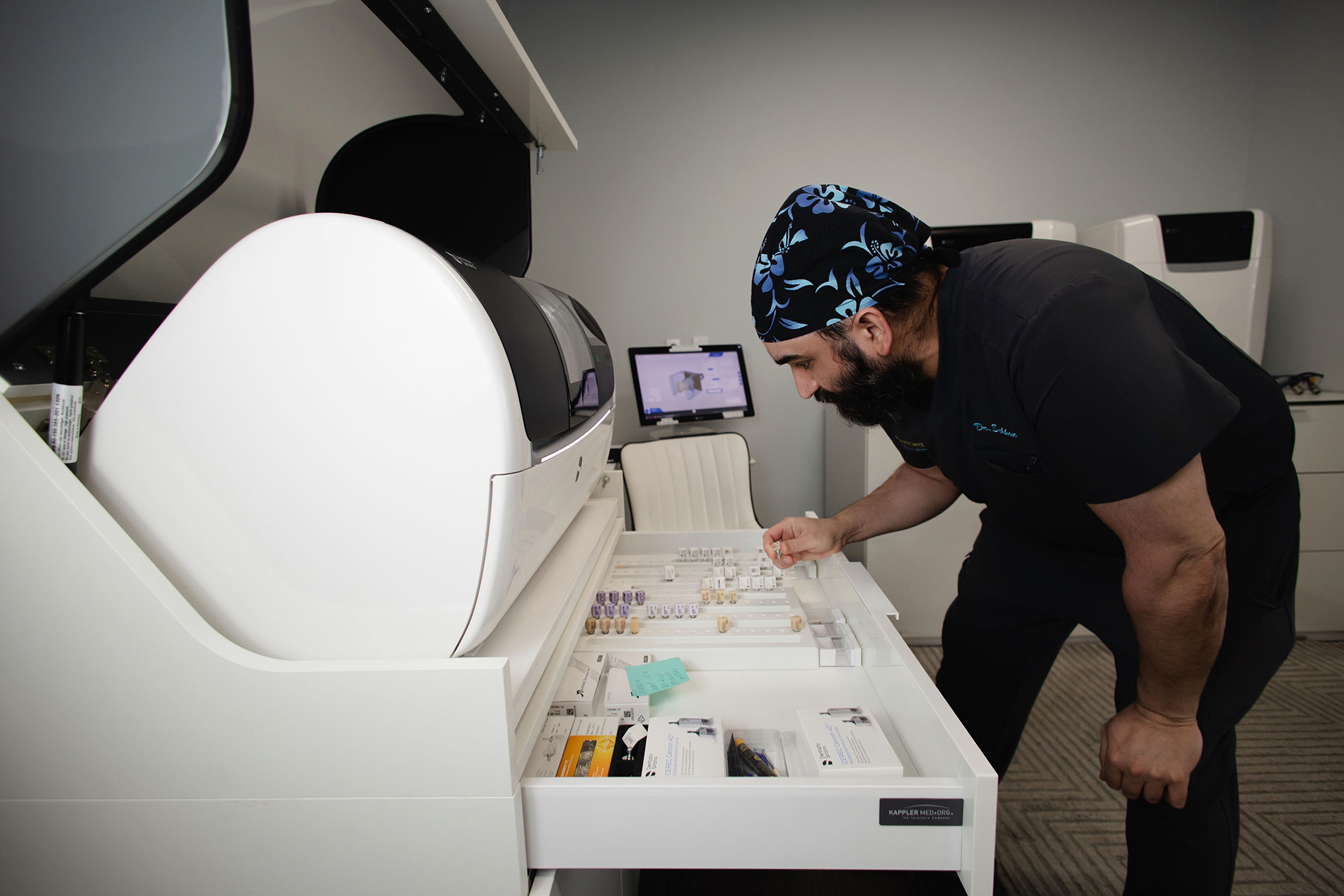 Man in medical scrubs and cap examining dental or medical components in an open equipment drawer.