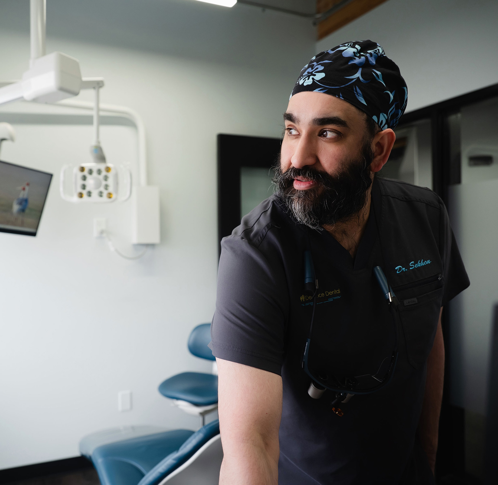 Bearded male dentist wearing black scrubs and cap in a dental office with equipment visible.