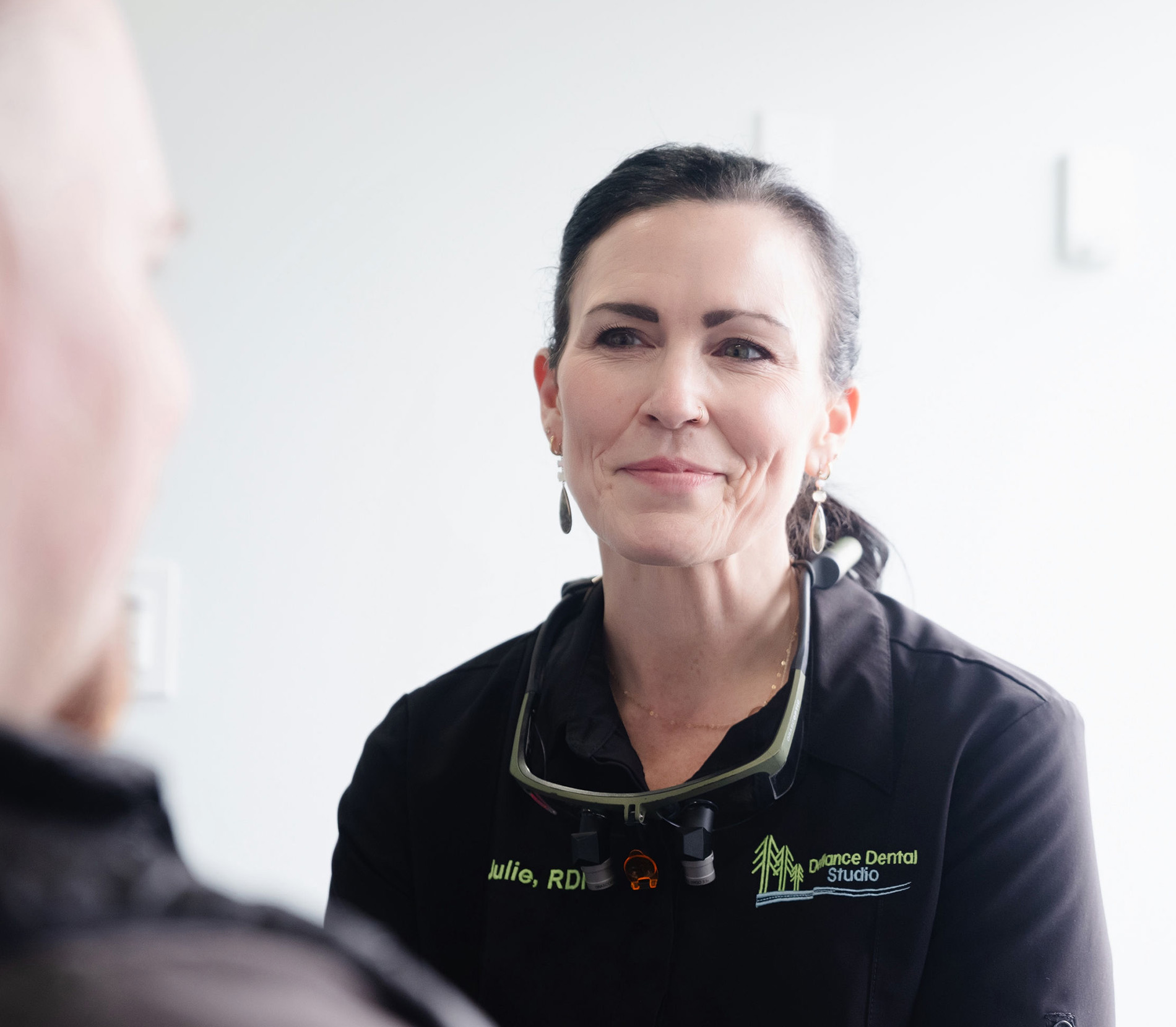 Female dental professional wearing a black uniform and magnifying glasses talking to a patient.