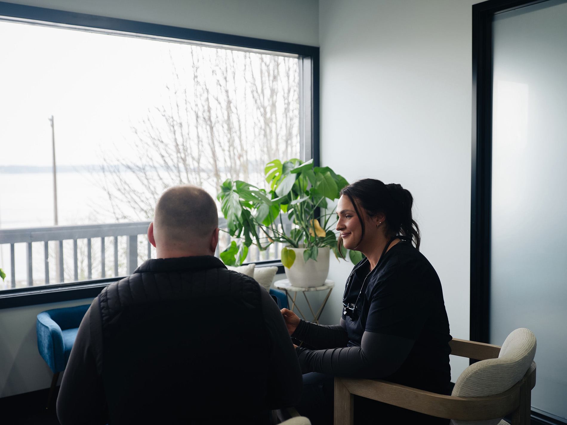 A woman in scrubs talking to a man in a vest in a bright room with a large window and plants.
