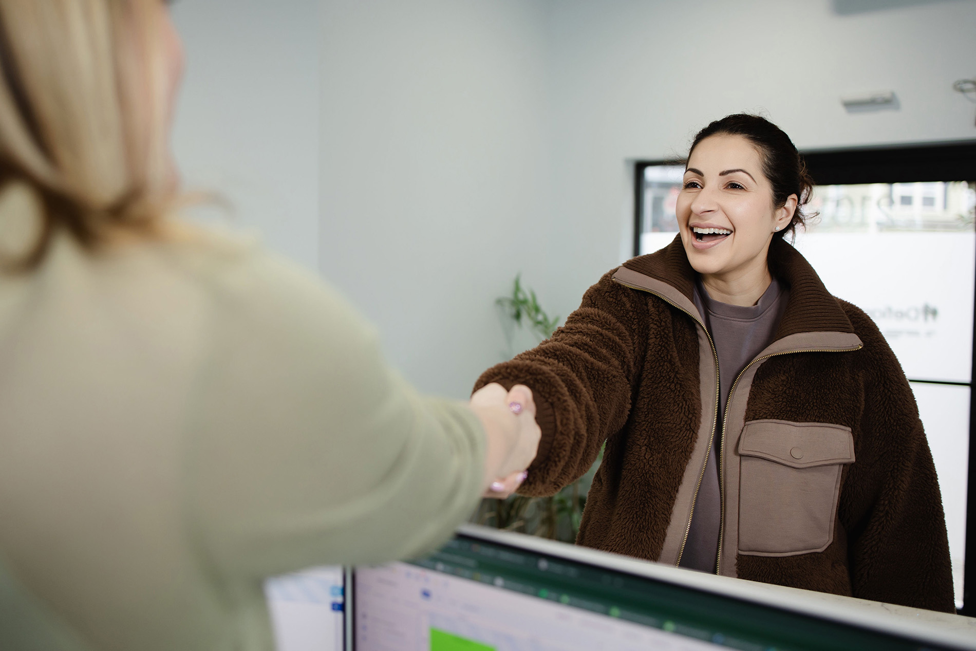 Smiling woman in brown jacket shakes hands with another person across a desk in an office.