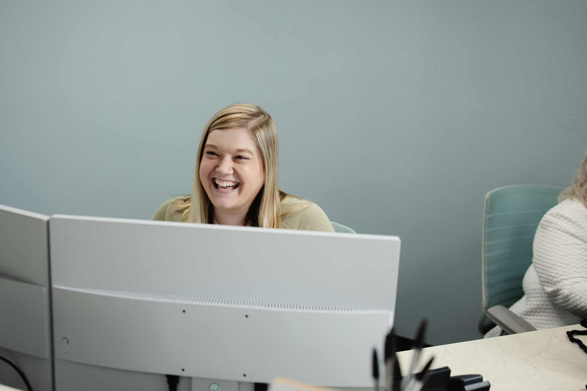 Smiling woman with blonde hair sitting behind dual computer monitors in an office.