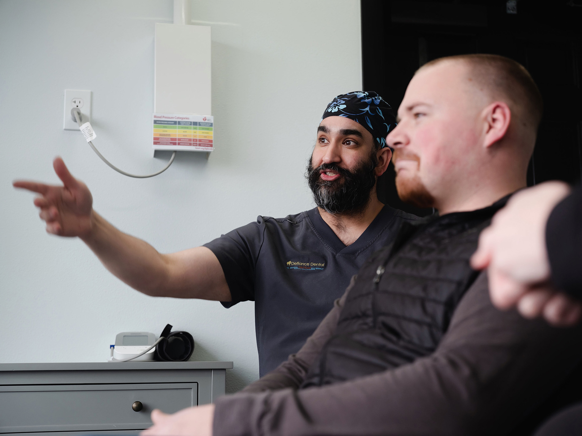 A bearded man in scrubs gestures while explaining something to a seated man in a black vest.