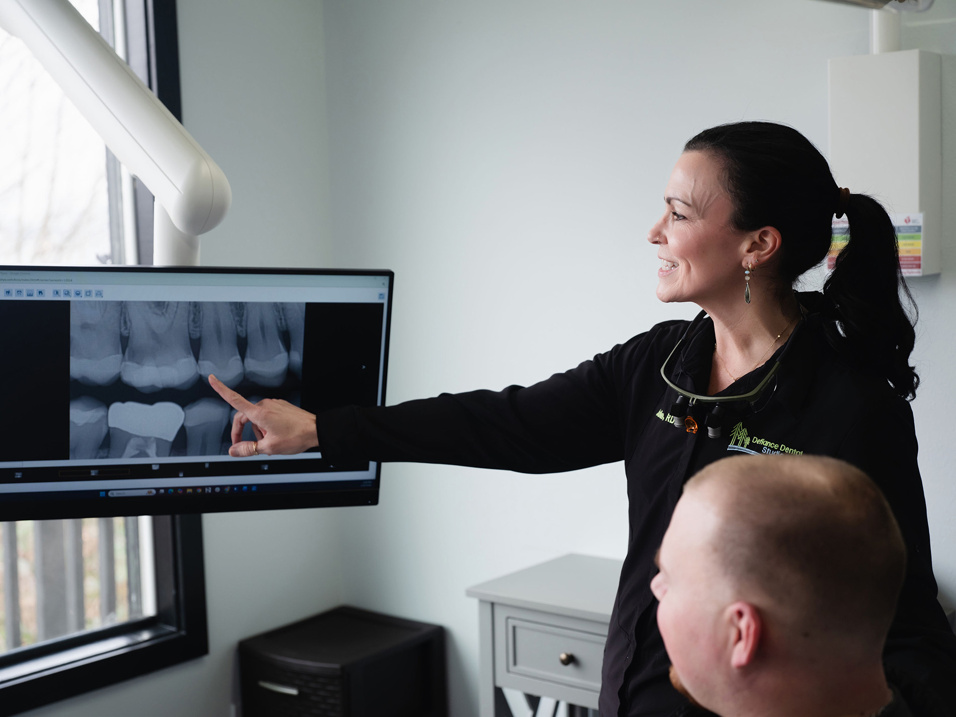 Dentist pointing at a dental X-ray on a screen while explaining to a seated patient in a clinic.