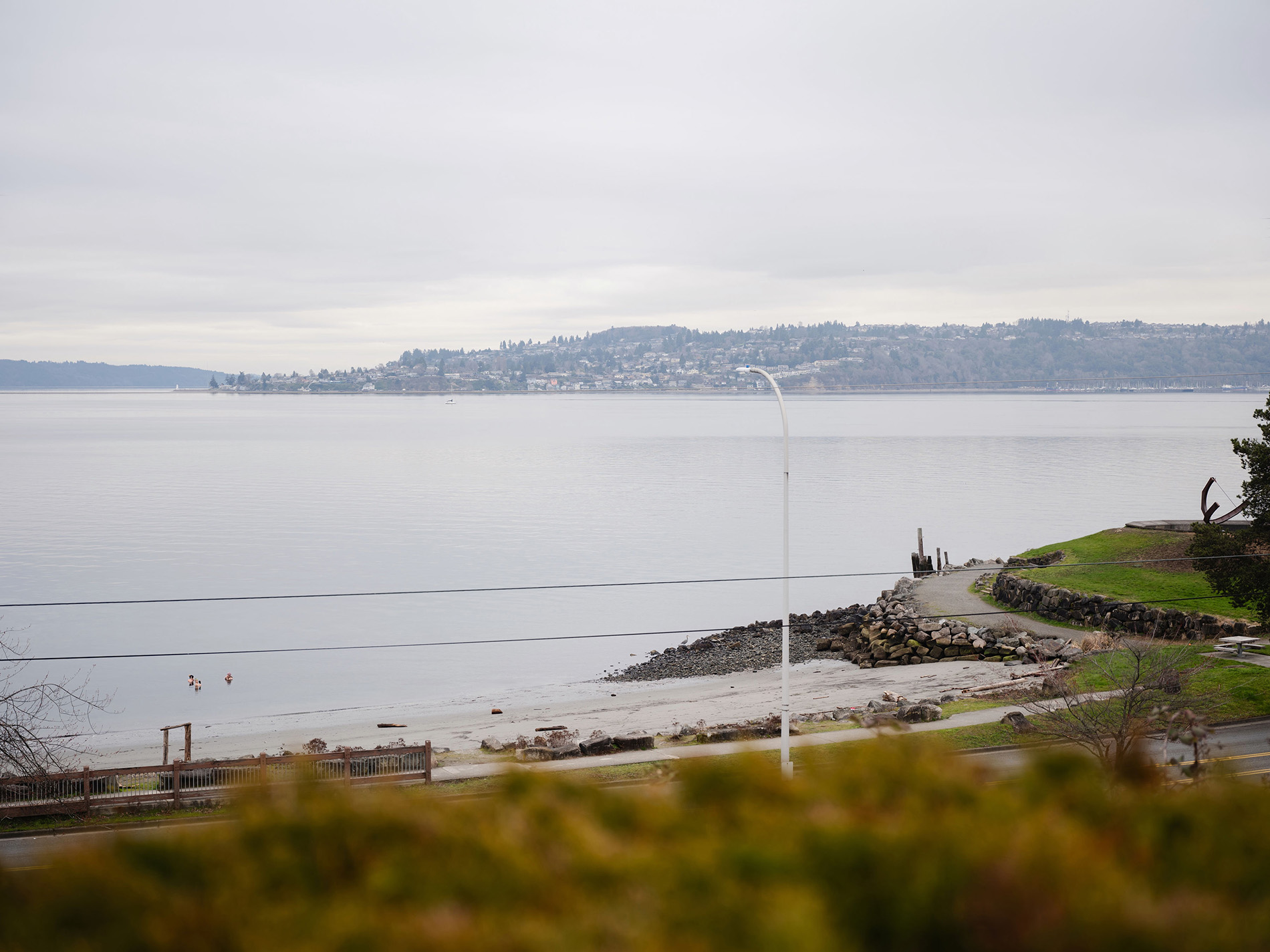 Calm shoreline with two people in water, rocky pier, and distant hillside with houses under gray sky.