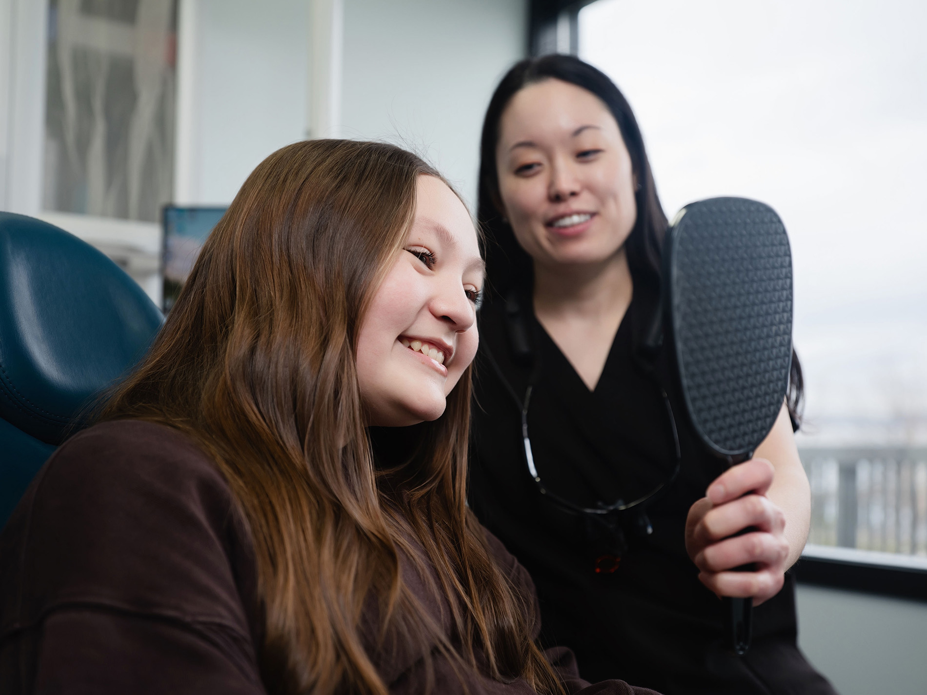 Dentist showing a smiling young girl her teeth reflection in a handheld mirror.
