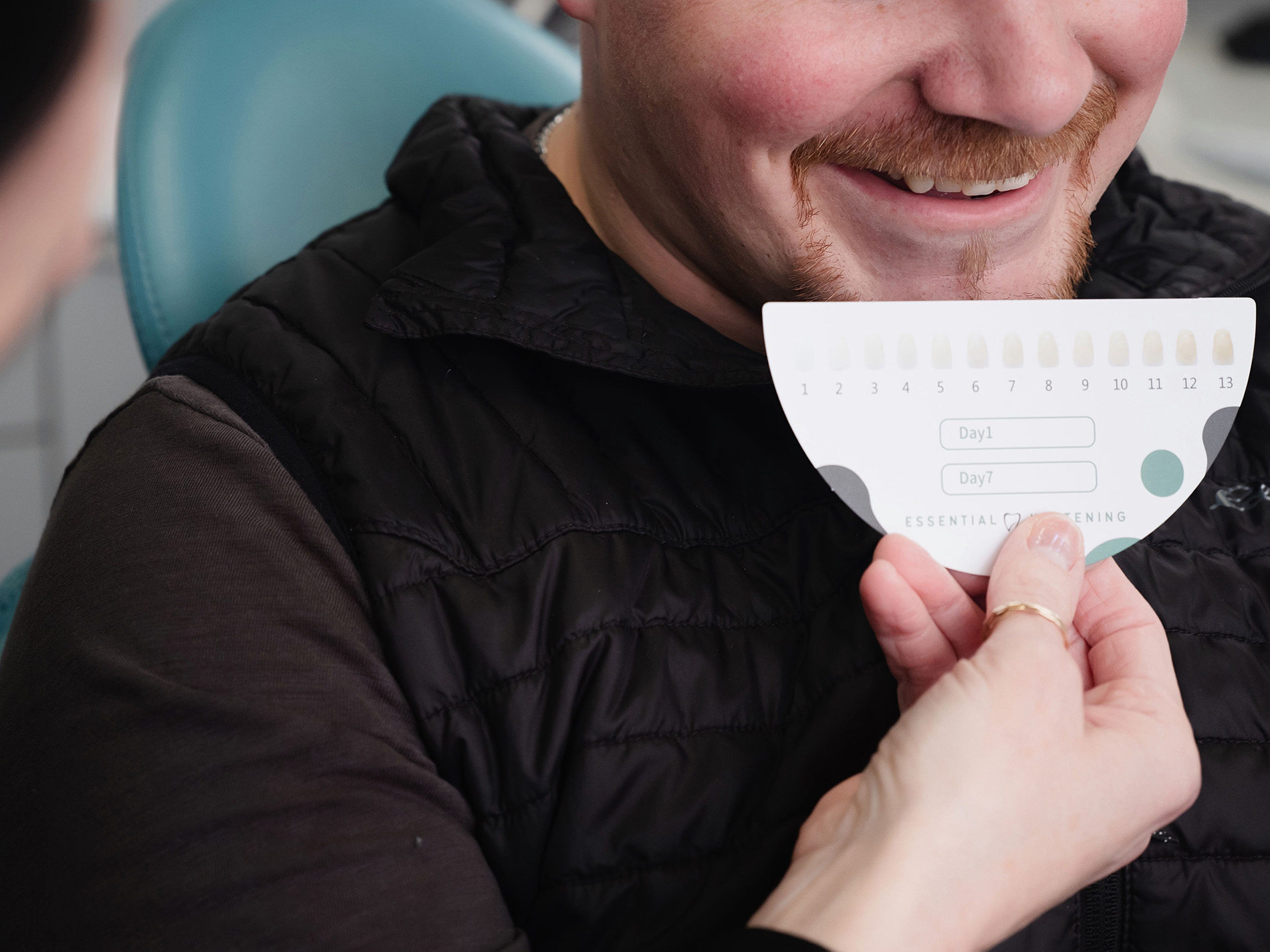 Man smiling as a dental professional compares his teeth to a shade guide card for whitening.