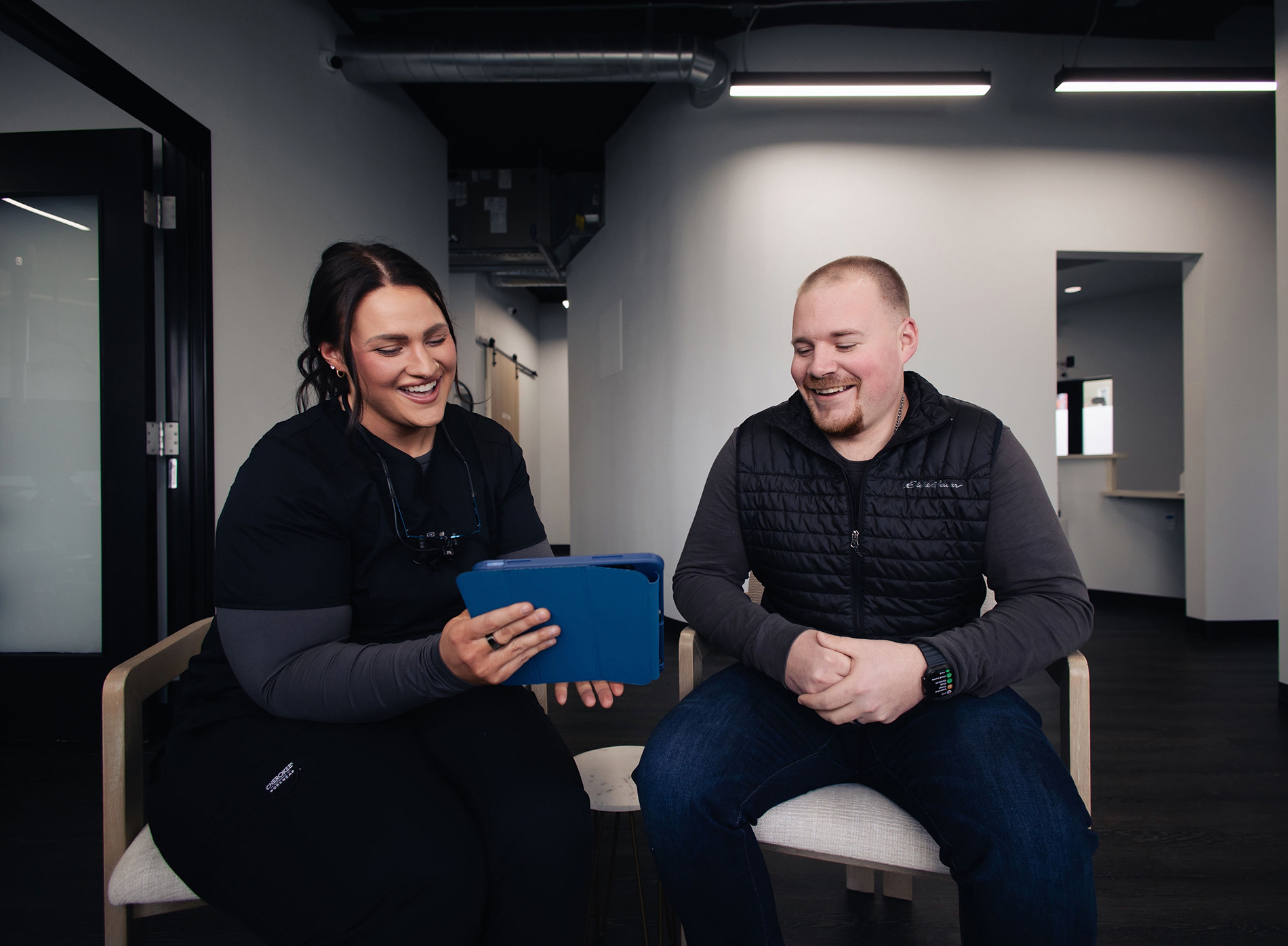 Dentist and male patient sitting and smiling while looking at a tablet in a modern office.