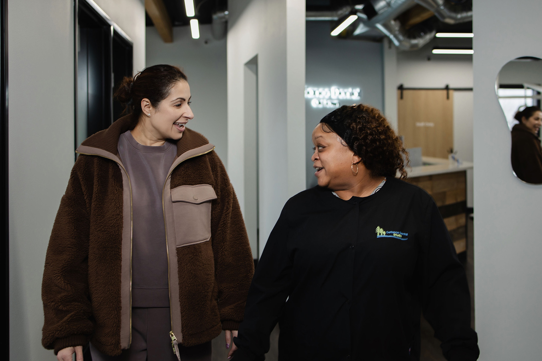 Two women smiling and talking while walking inside a modern dental office hallway.
