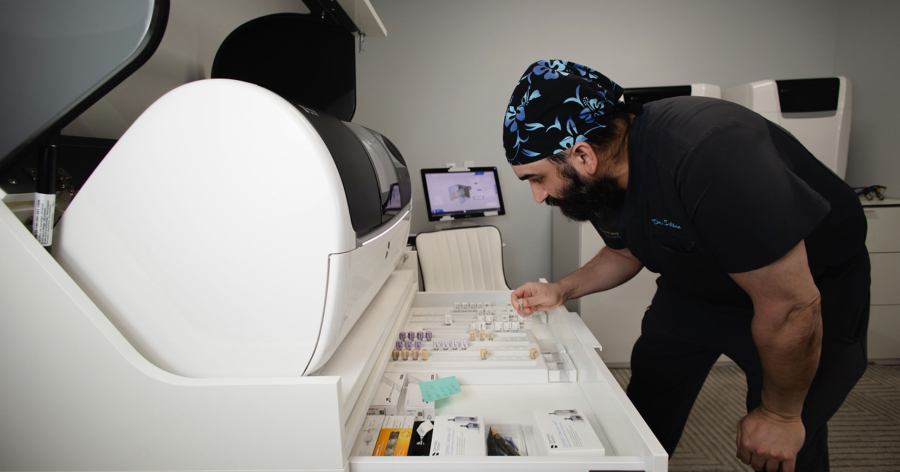 Man in black scrubs and floral cap sorting dental crown samples beside a large white dental scanner.
