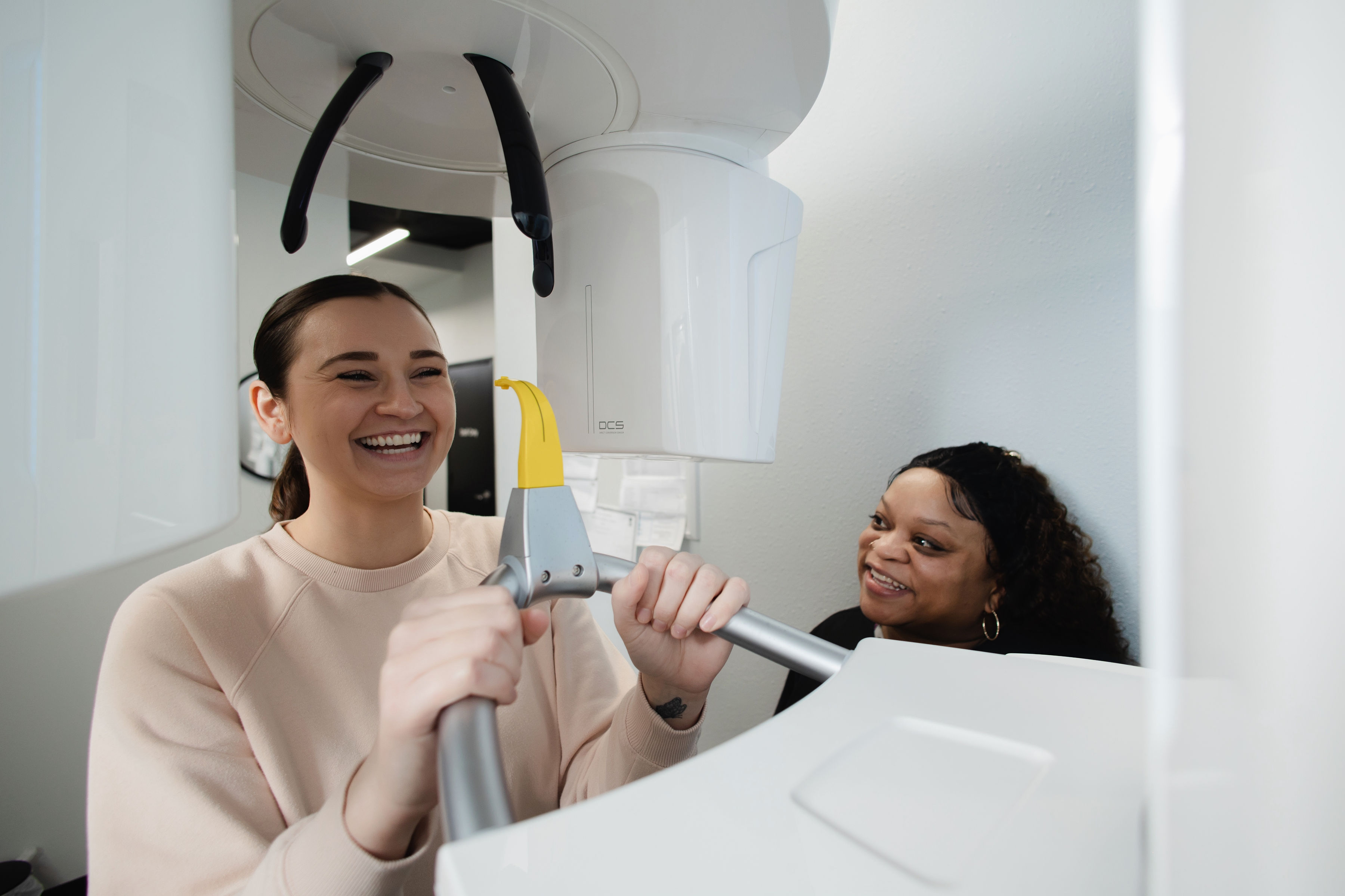 Smiling woman holding handles of a dental 3D imaging machine while a technician watches.