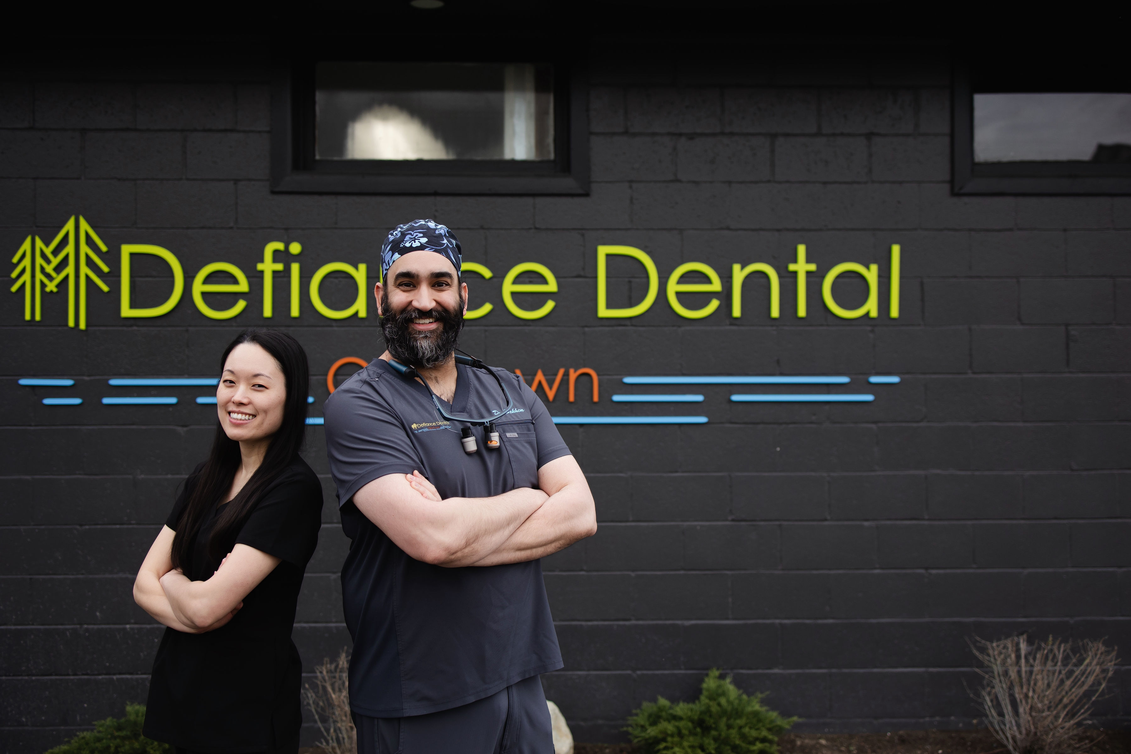 Two smiling dental professionals standing with arms crossed in front of Defiance Dental building.