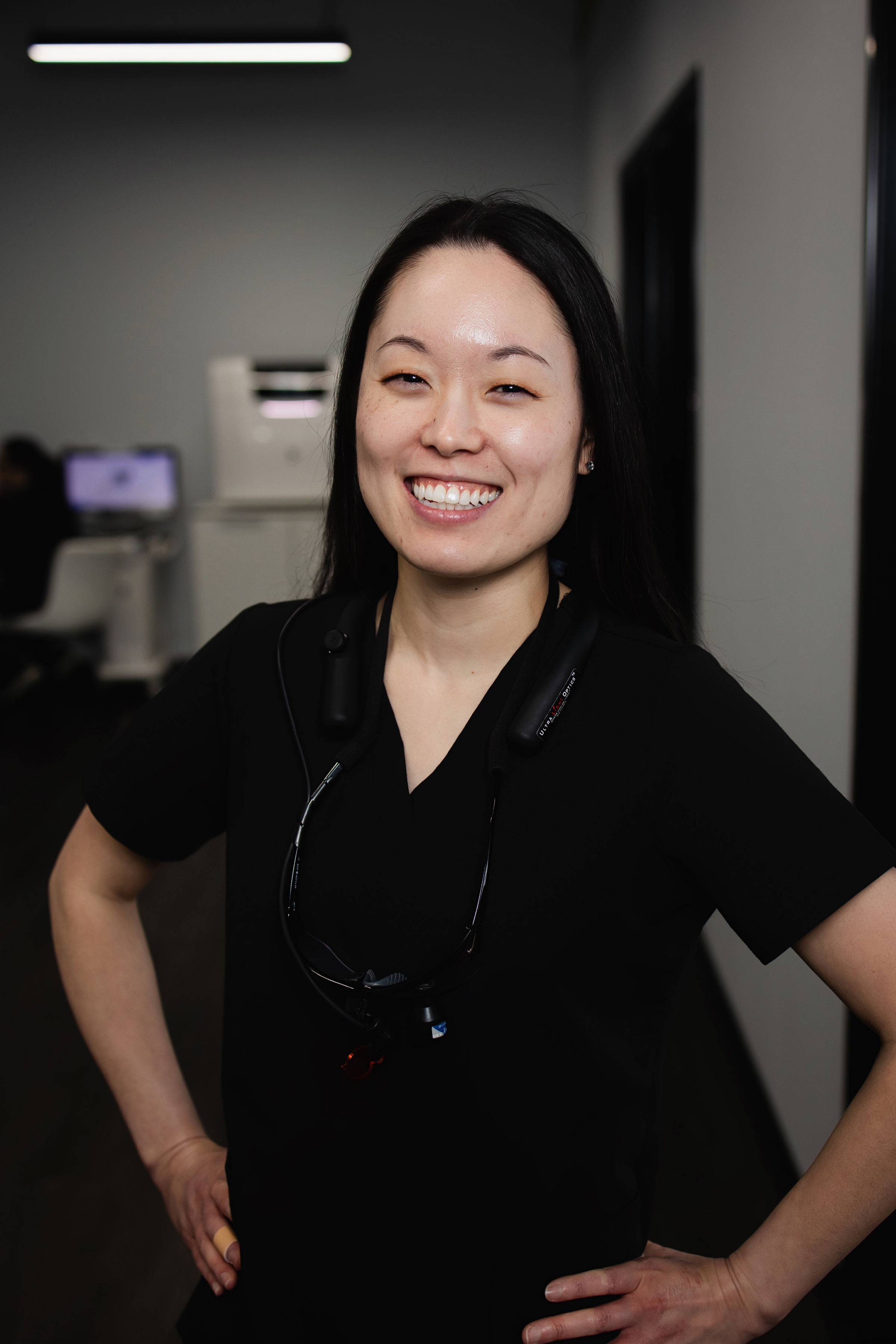 Smiling woman in black scrubs wearing dental loupes around her neck in a clinic hallway.