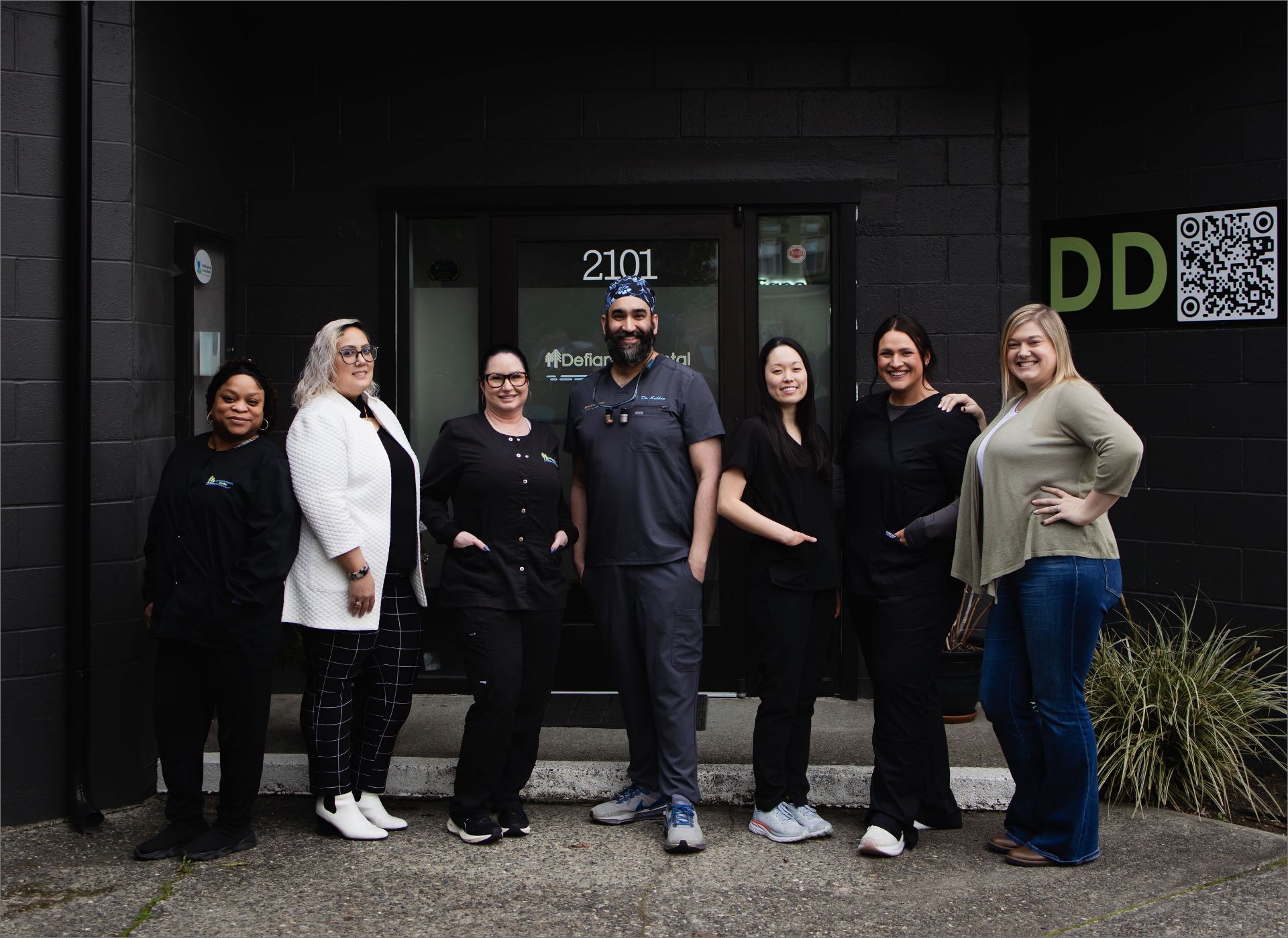 Seven dental professionals standing and smiling outside a clinic entrance with address 2101.