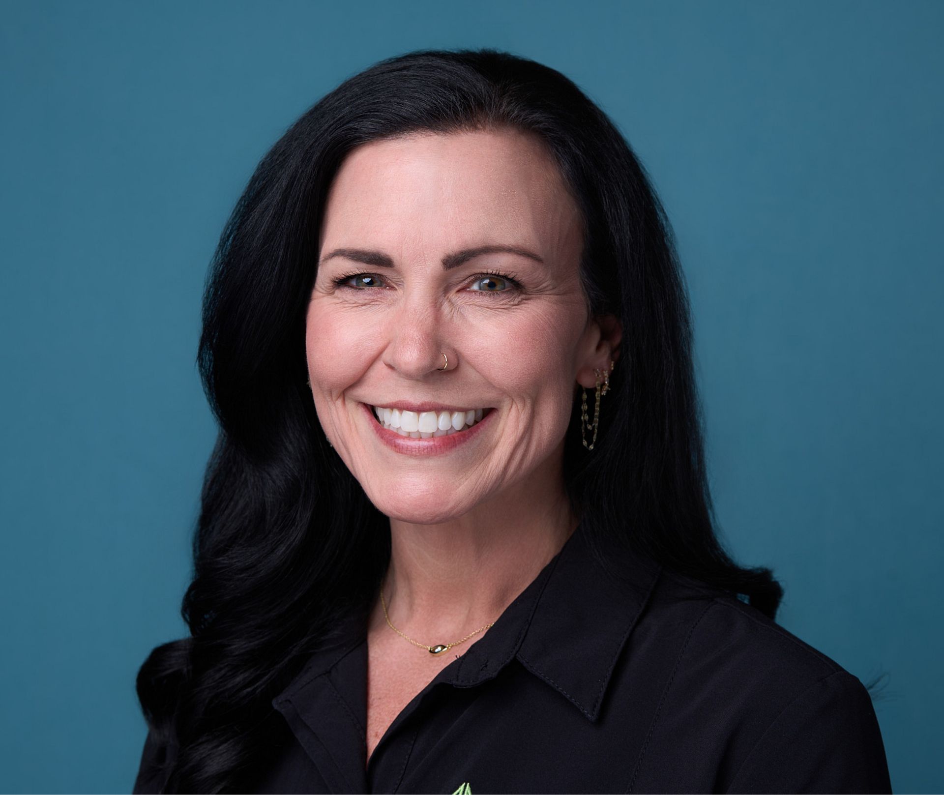 Smiling woman with long black hair, wearing a black collared shirt and gold jewelry against a blue background.