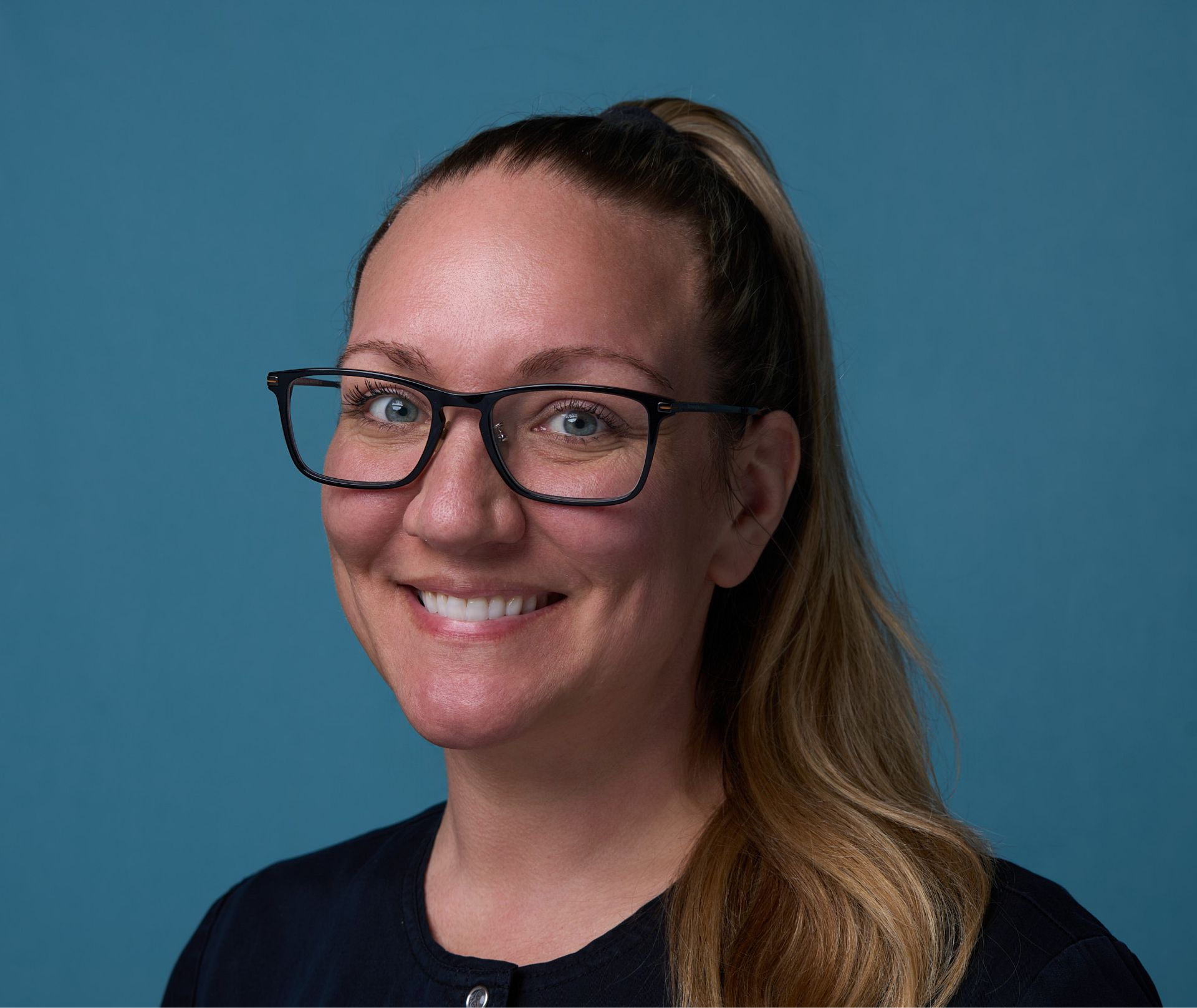 Smiling woman with glasses and blond hair tied back against a blue background.