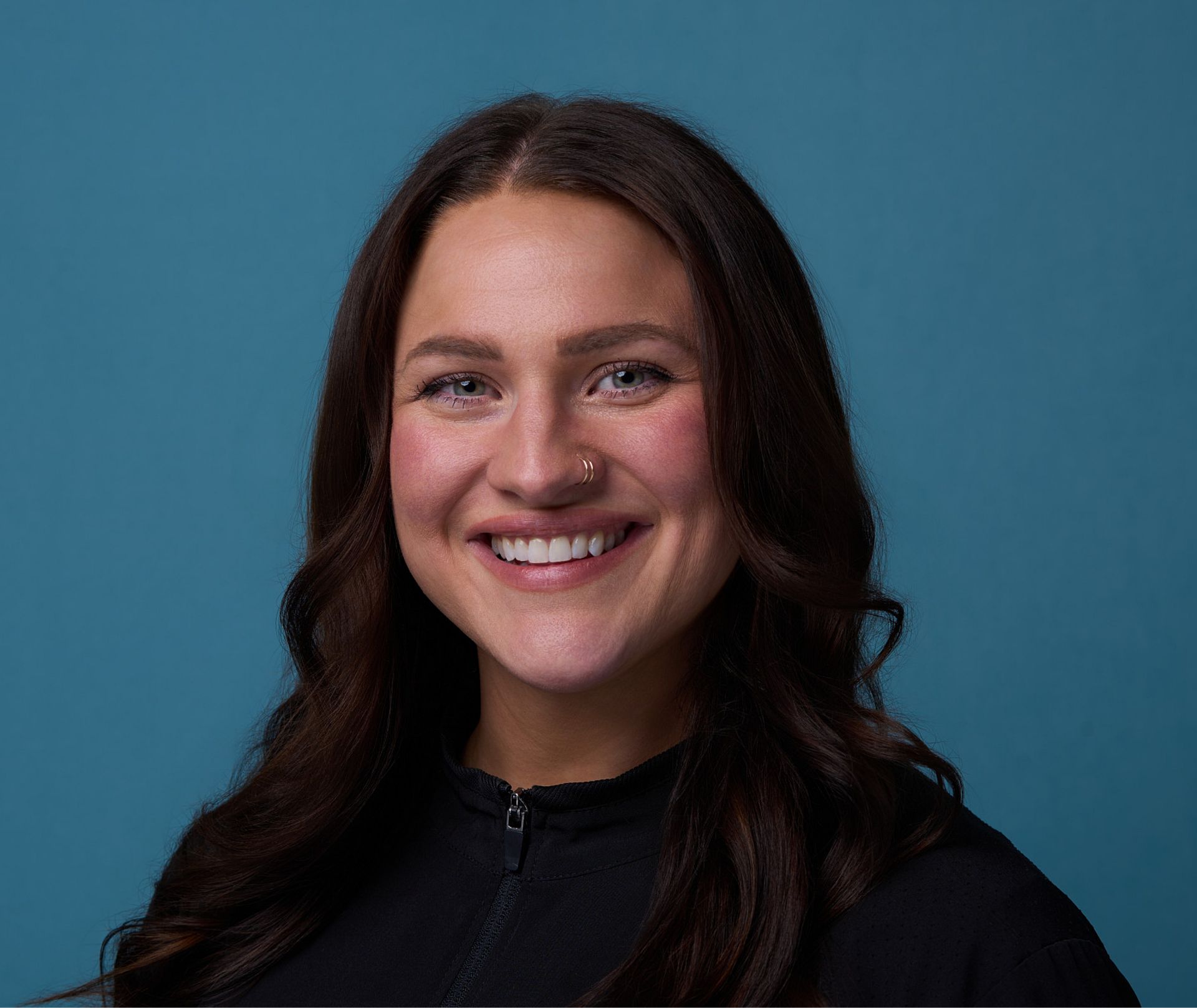 Smiling woman with long dark hair and nose ring wearing black jacket against blue background.