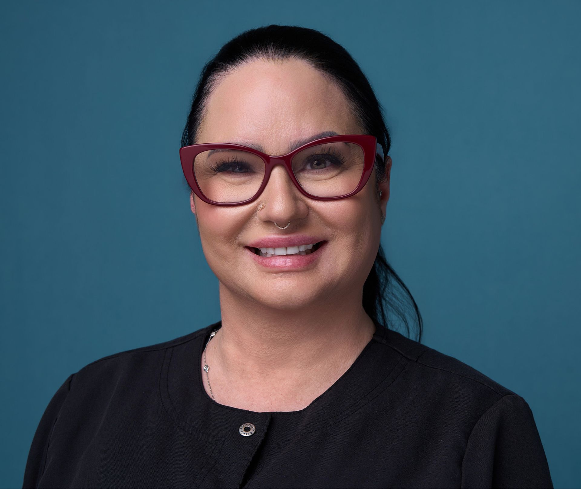 Smiling woman with dark hair, red glasses, nose ring, and black top against a blue background.