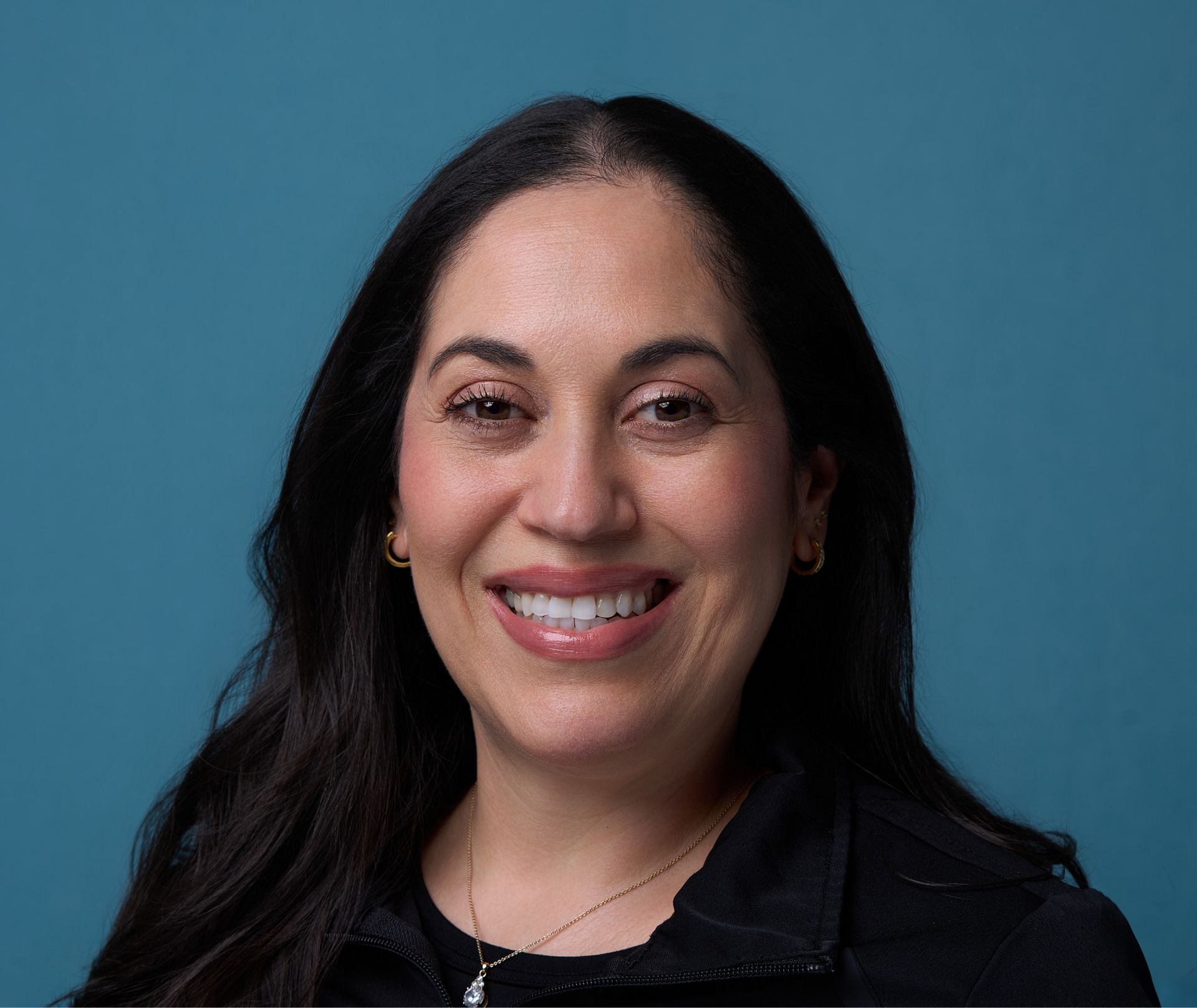 Smiling woman with long dark hair, wearing a black top, gold earrings, and a pendant necklace.