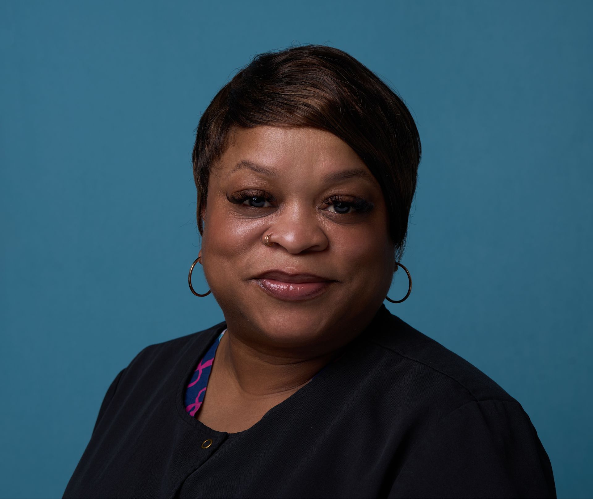 Smiling woman with short hair, hoop earrings, and nose ring against a blue background.