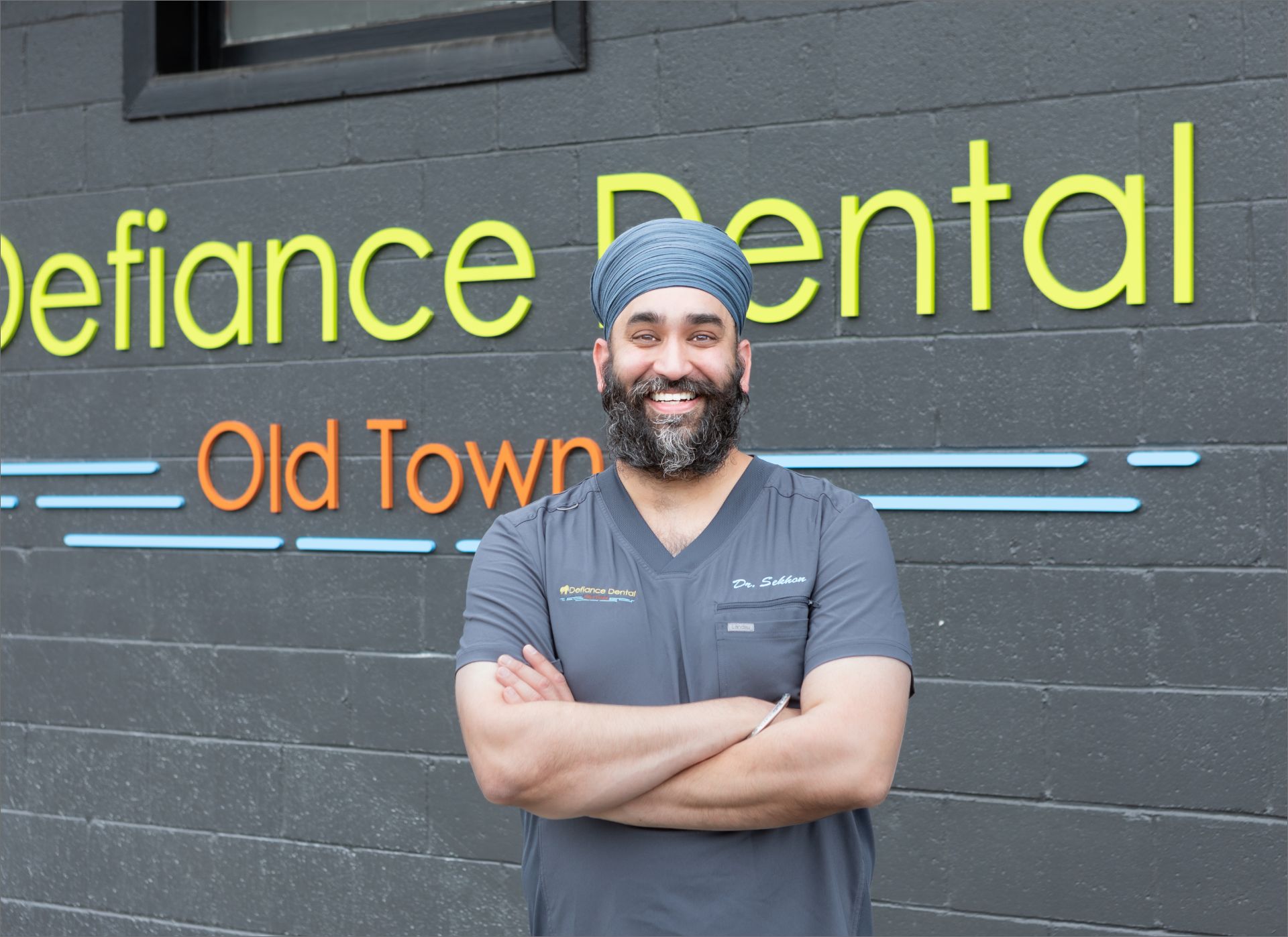 Smiling man with turban and beard in gray scrubs stands with arms crossed outside Defiance Dental Old Town.