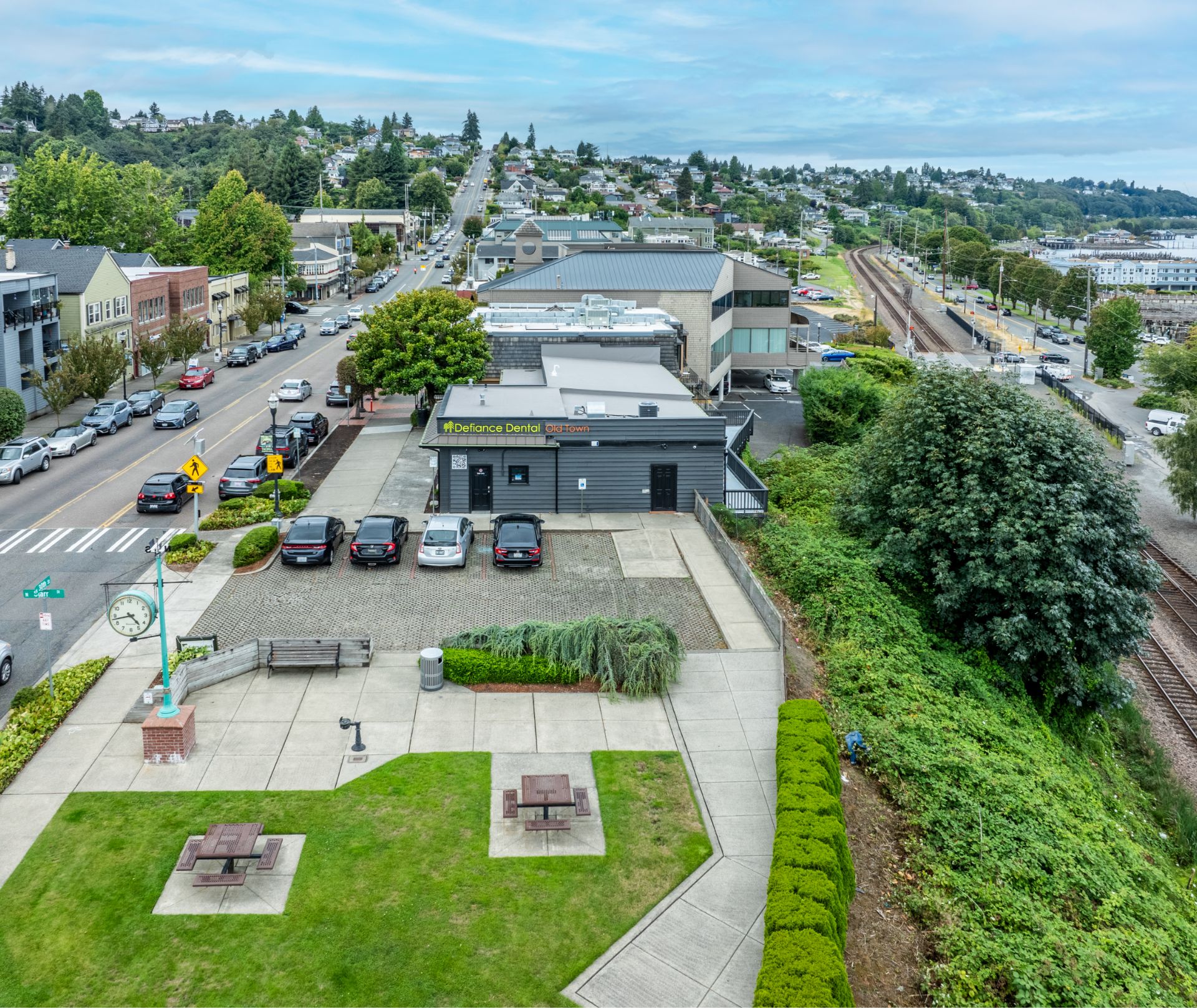 Aerial view of a town street, dental office, parking lot, park benches, railway tracks, and hillside houses.