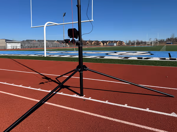 Tripod standing on a red running track near a football field with goalposts and clear blue sky.
