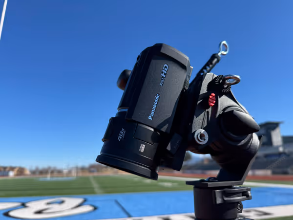 Close-up of a Panasonic Full HD camcorder mounted on a tripod with a sports field and blue sky in the background.