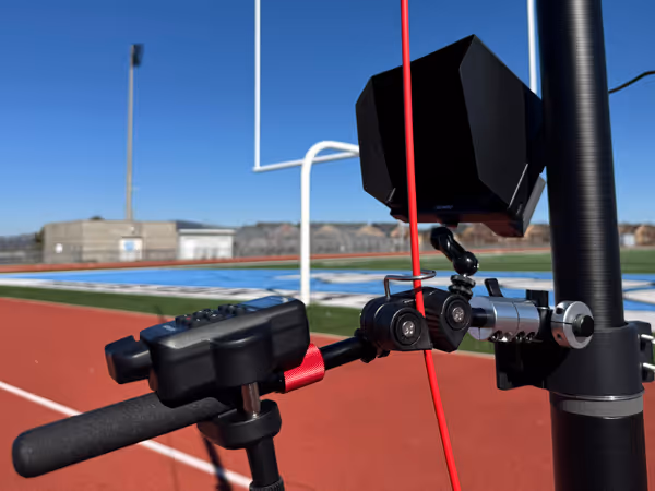 Close-up of a pan and tilt arm camera mount attached to a pole on a sports field with a running track and football goalpost in the background.