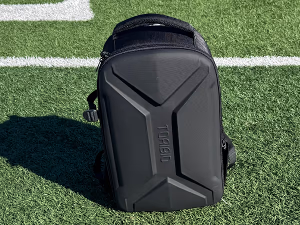 Black Torba hard-shell backpack standing on green artificial turf with white field markings in sunlight.