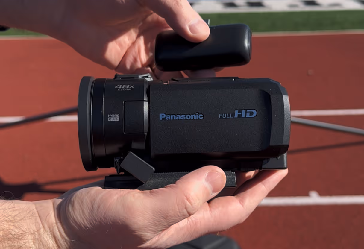 Hands holding a Panasonic Full HD camcorder over a red outdoor sports track.