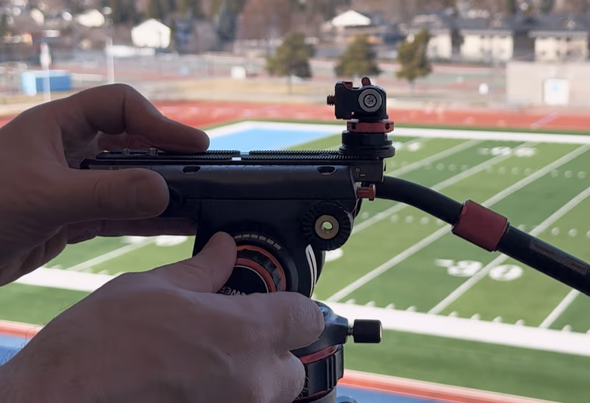 Close-up of hands adjusting a fluid head tripod with a sports field in the background.