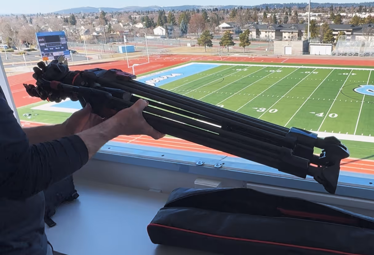 Person holding a folded heavy-duty tripod inside a press box overlooking a football field and running track.