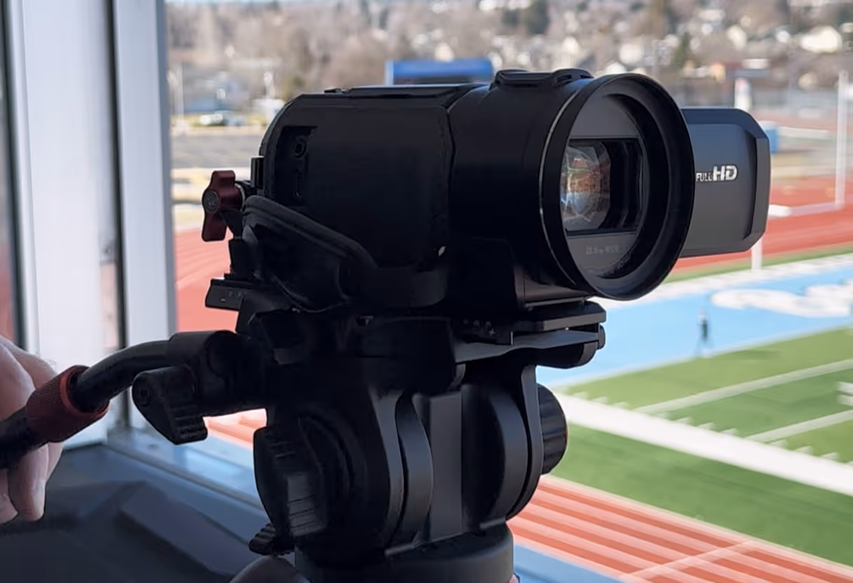 Black Full HD video camera mounted on a tripod filming a sports field with a running track.
