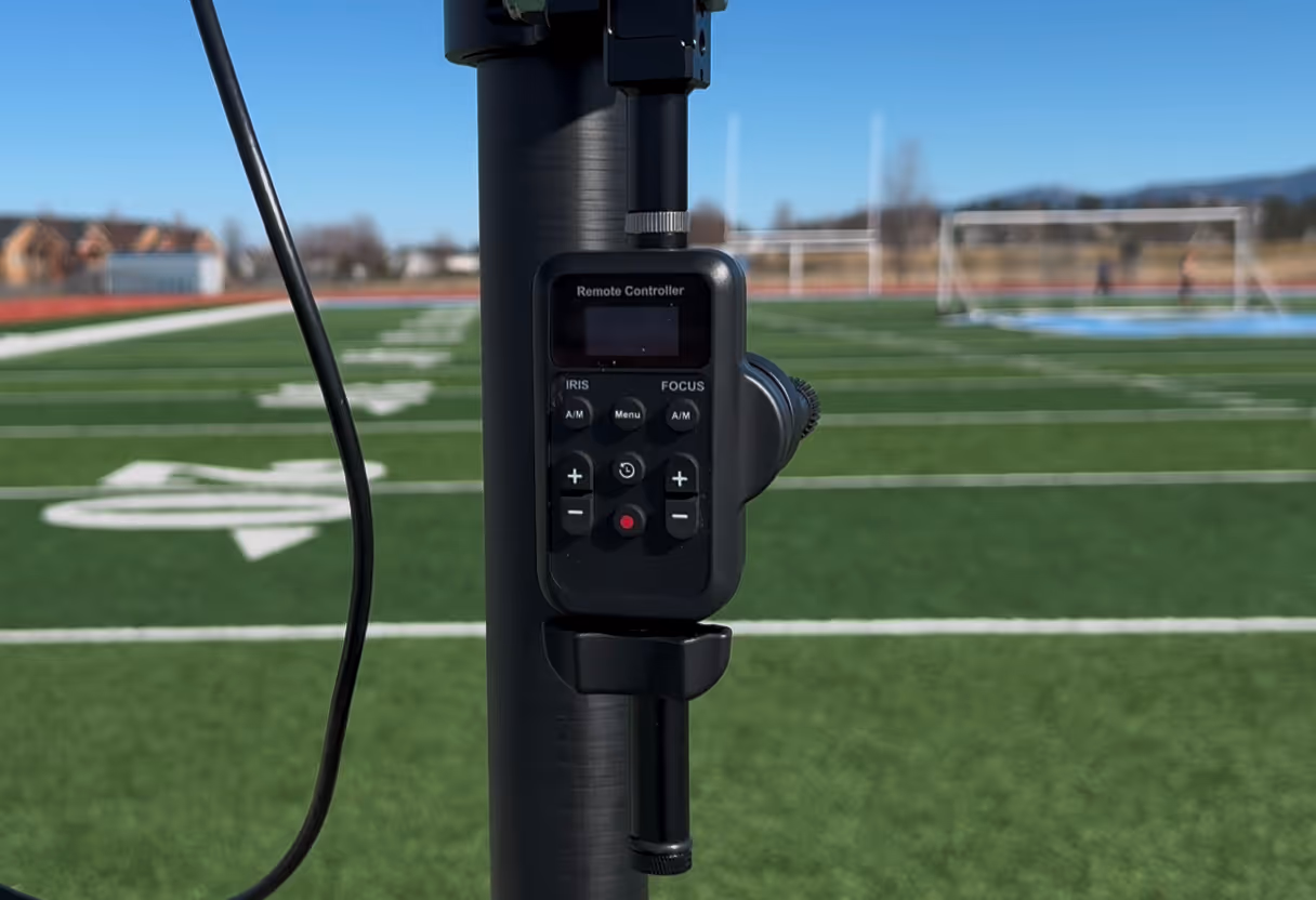 Close-up of a black remote controller mounted on a pole with a green sports field and soccer goals in the background.