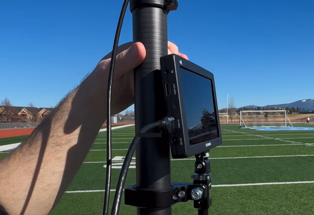 Hand holding a black monitor mounted on a pole on a sports field with clear blue sky.