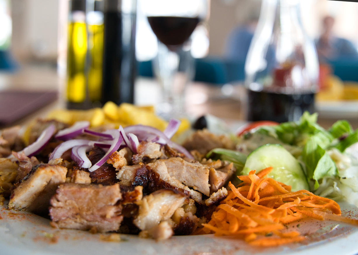 Teller mit gegrilltem Fleisch, roten Zwiebeln, geraspelten Karotten, Salat, Gurken und Pommes Frites, verschwommener Hintergrund mit Weinflaschen und Weinglas.