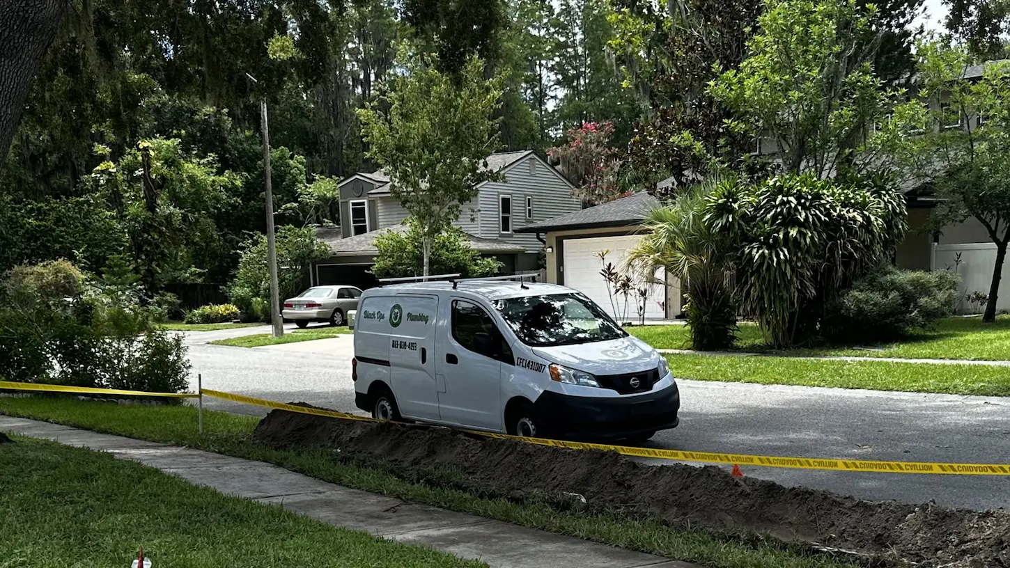 White Nissan work van parked by a roadside excavation with yellow caution tape in a residential neighborhood with green trees and houses.