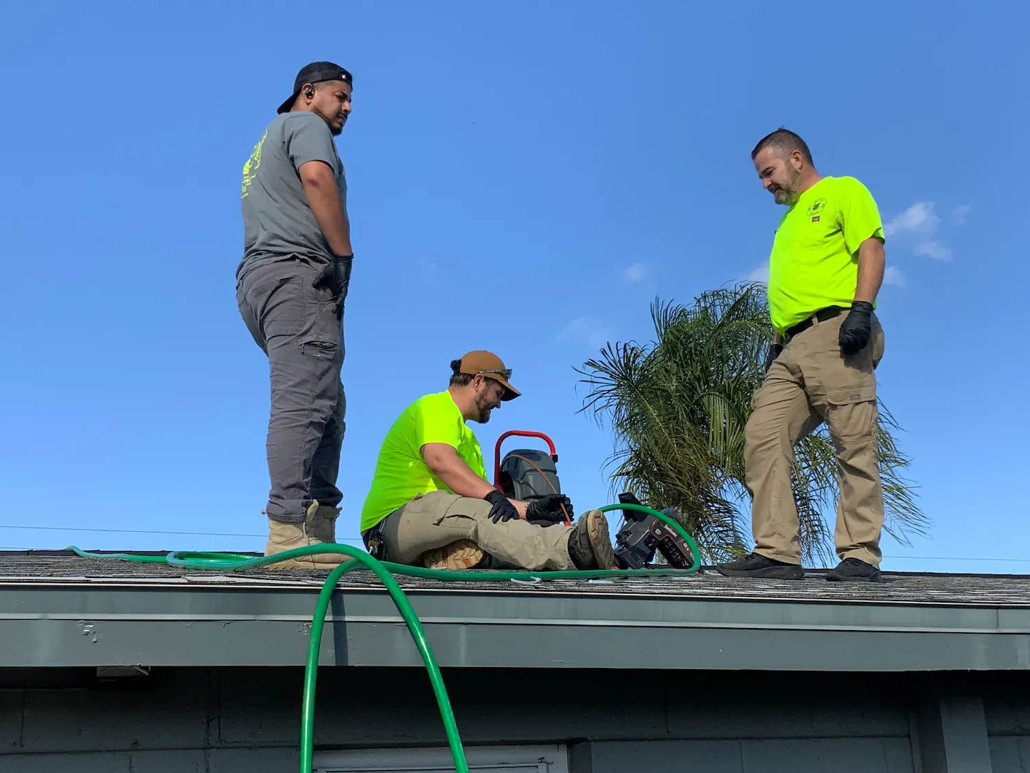 Three workers on a roof under a clear blue sky, with two standing and one sitting operating equipment connected to green hoses.